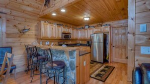 L’intérieur d’un chalet en bois comprend un bar central en pierre, des meubles de rangement en bois, un réfrigérateur en acier inoxydable et un mur en pierre.