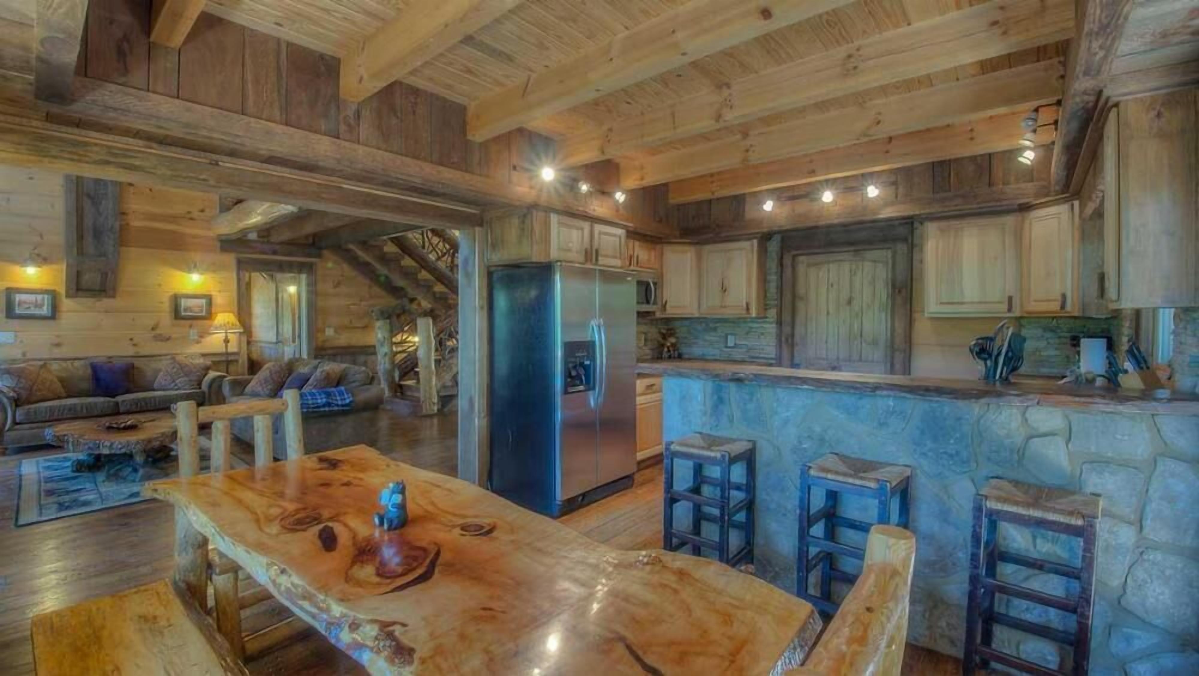 Interior d'una cabana rústica amb taula de dinar de fusta, tamborets de bar, cuina amb encimera de pedra i zona d'estar acollidora amb sofà i llar de foc.