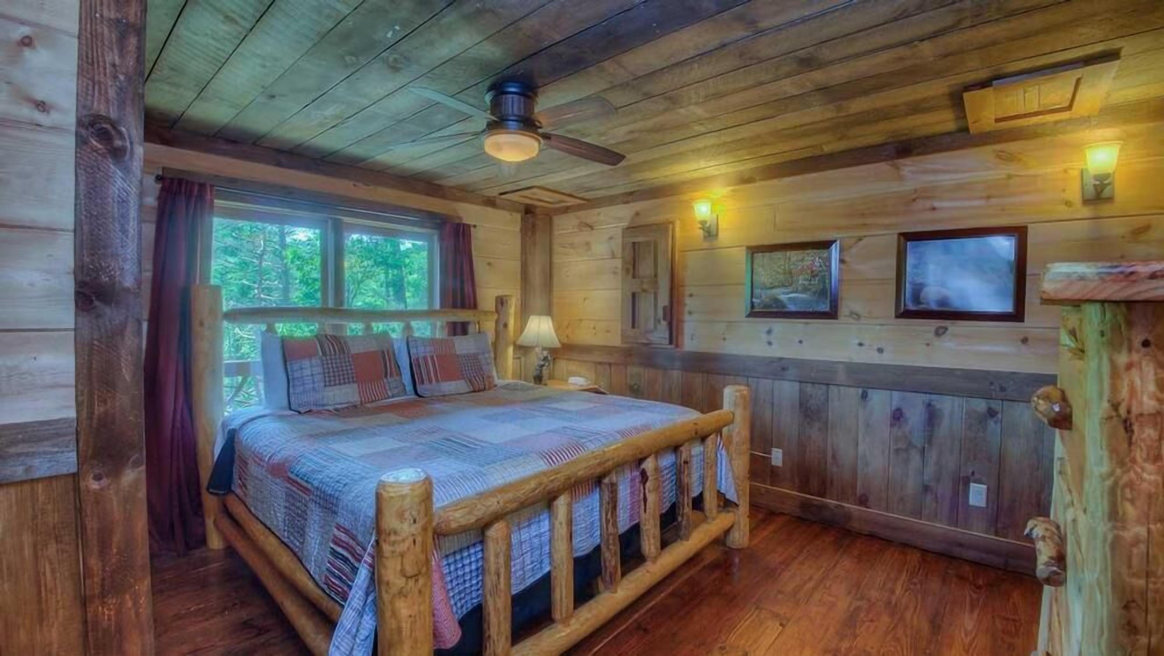 Une chambre dans une cabane en bois, avec un lit, un ventilateur de plafond et une fenêtre donnant sur la verdure.