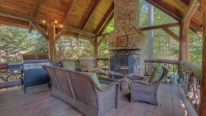 An outdoor covered patio with a stone fireplace, wicker furniture, and a grill. - Whitewater Wilderness- Riverside Retreat | Outdoor Living | Luxury Kitchen (Blue Ridge)