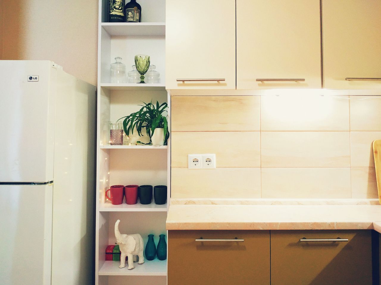 Une cuisine avec des meubles blancs, un réfrigérateur blanc et des étagères où sont disposés divers objets, notamment des tasses, une plante et des objets décoratifs.