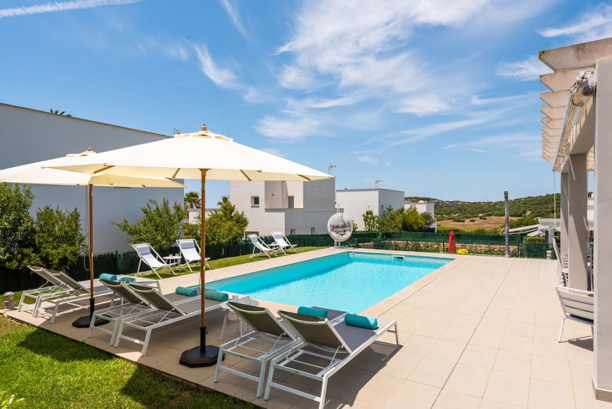 Piscina com espreguiçadeiras e guarda-sóis, cercada por um jardim e um edifício branco.