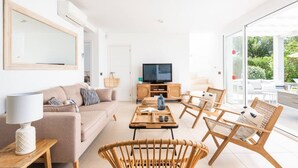 A modern living room with a sofa, coffee table, and wicker chairs. A television is on a wooden cabinet, and a large mirror hangs on the wall.