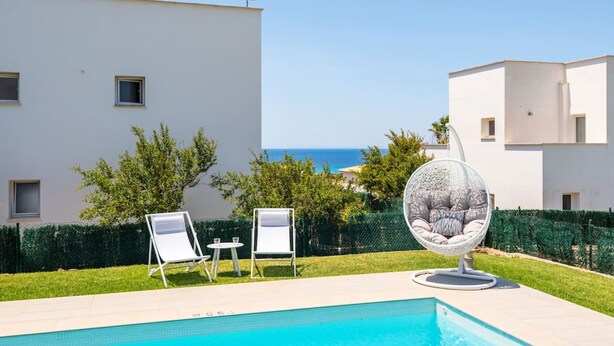 Ein Poolbereich mit zwei Liegestühlen und einem Kugelstuhl, umgeben von einem weißen Gebäude mit Blick auf das Meer.