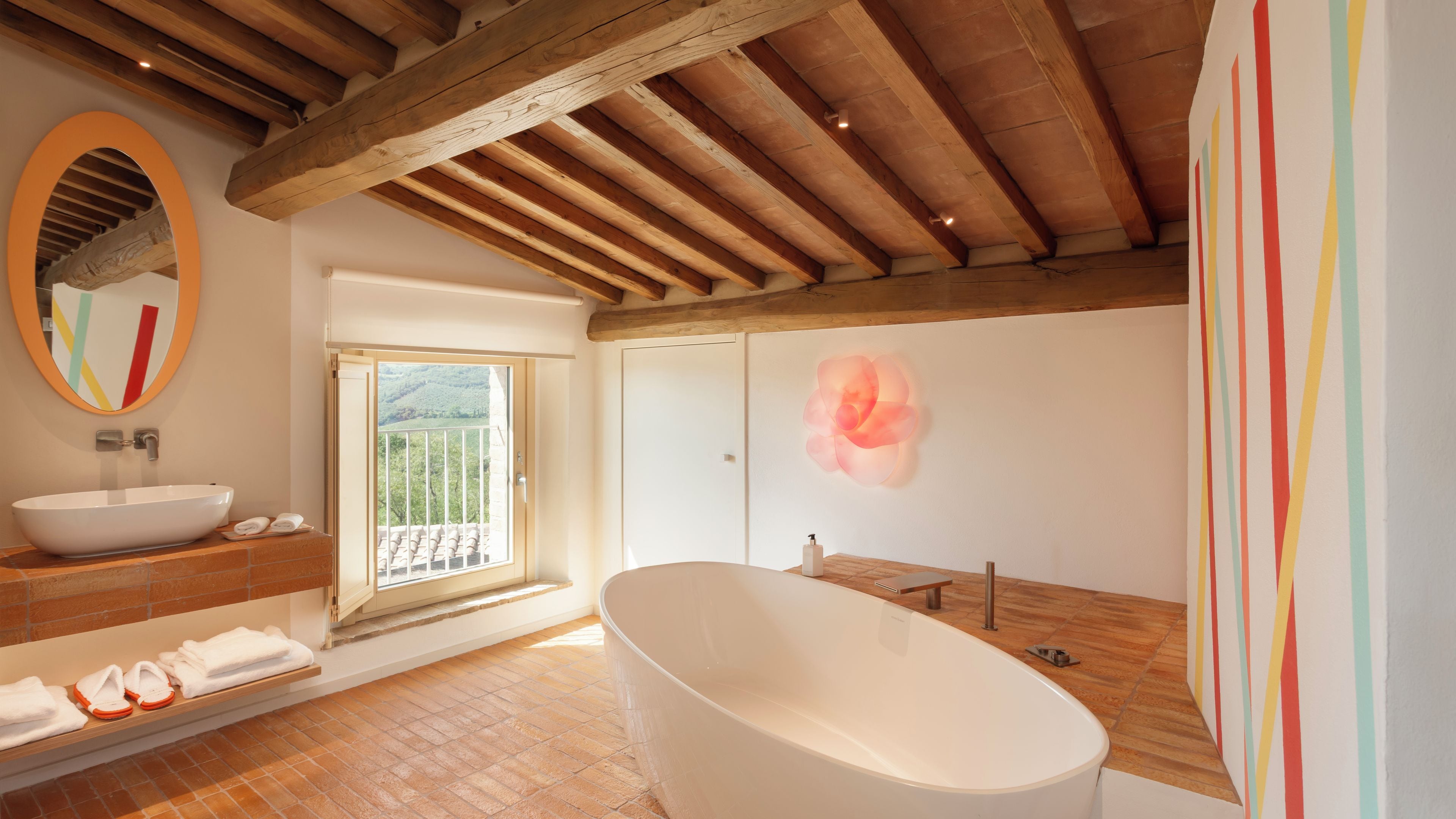 Un baño con una bañera independiente, un espejo grande y una ventana con vistas a una colina.
