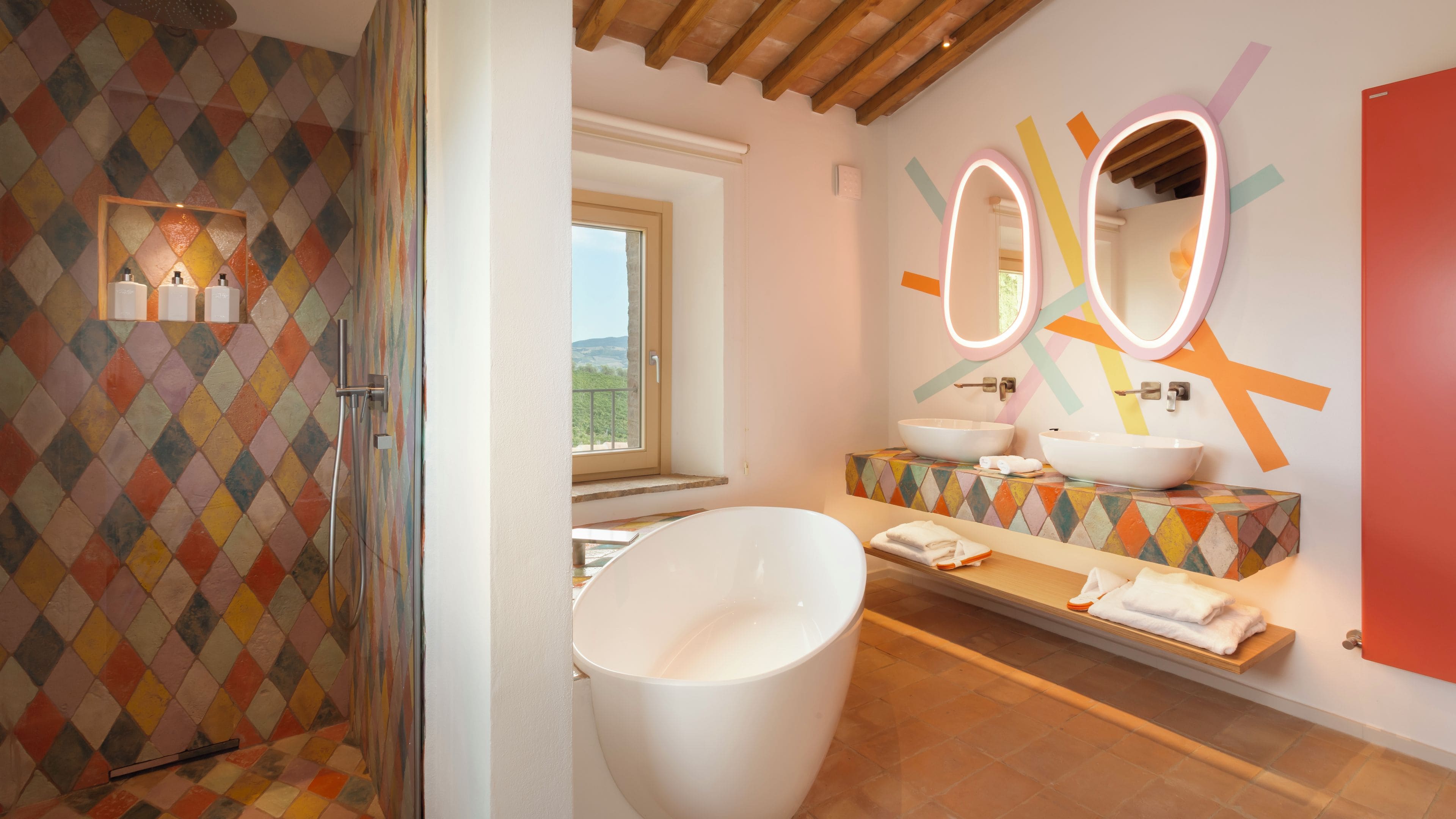 A modern bathroom with a freestanding bathtub, two sinks with round mirrors, and a colorful diamond-patterned shower wall.