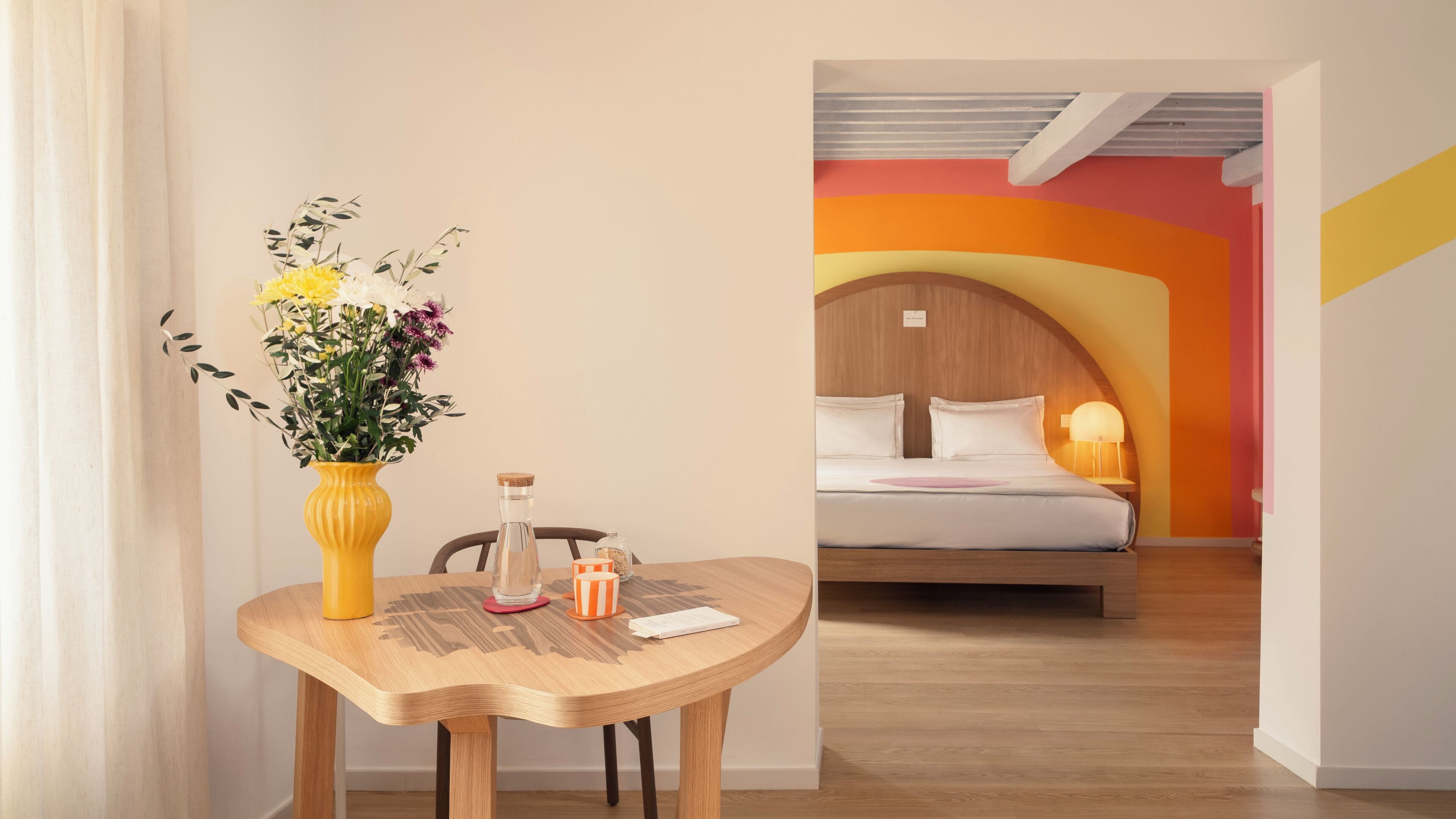 Una habitación con una mesa de madera, un florero con flores, una cama con cabecera y una lámpara.