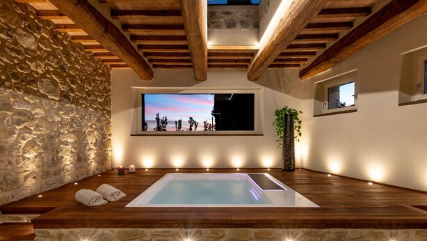 A spa area with a stone wall, wooden beams, a large window with a view, and a hot tub. - Mastrojanni Relais - Small Luxury Hotels of the World (Montalcino)