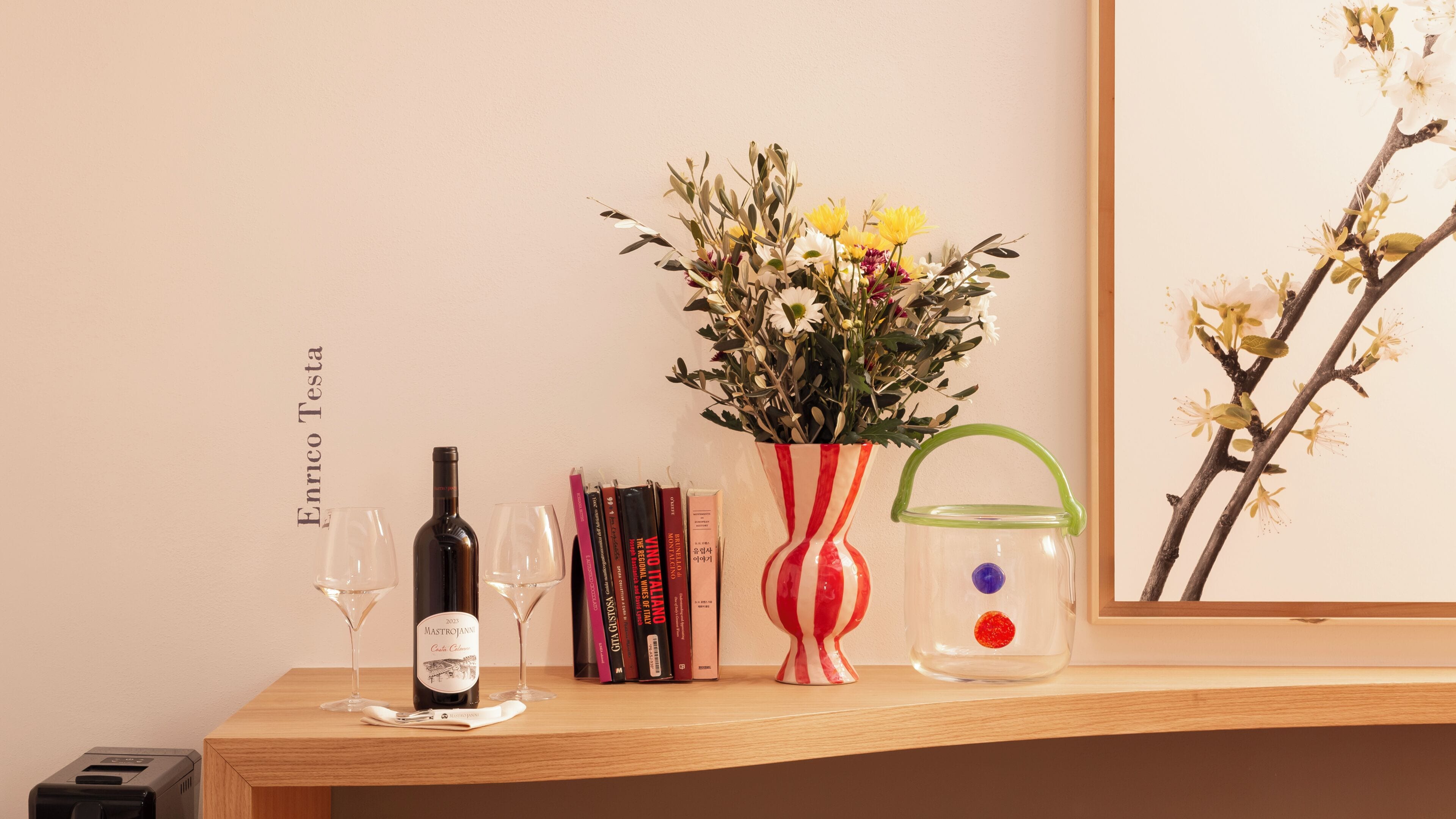A wooden shelf with a wine bottle, glasses, books, a striped vase with flowers, and a small decorative item.