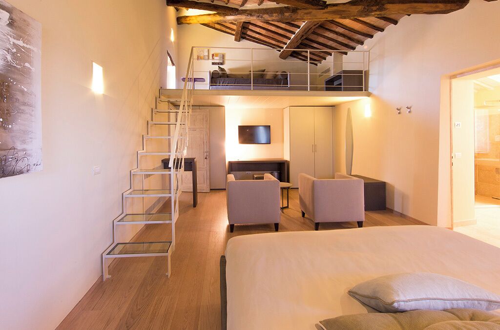 A loft-style bedroom with a bed, a desk, two chairs, a TV, and a staircase.