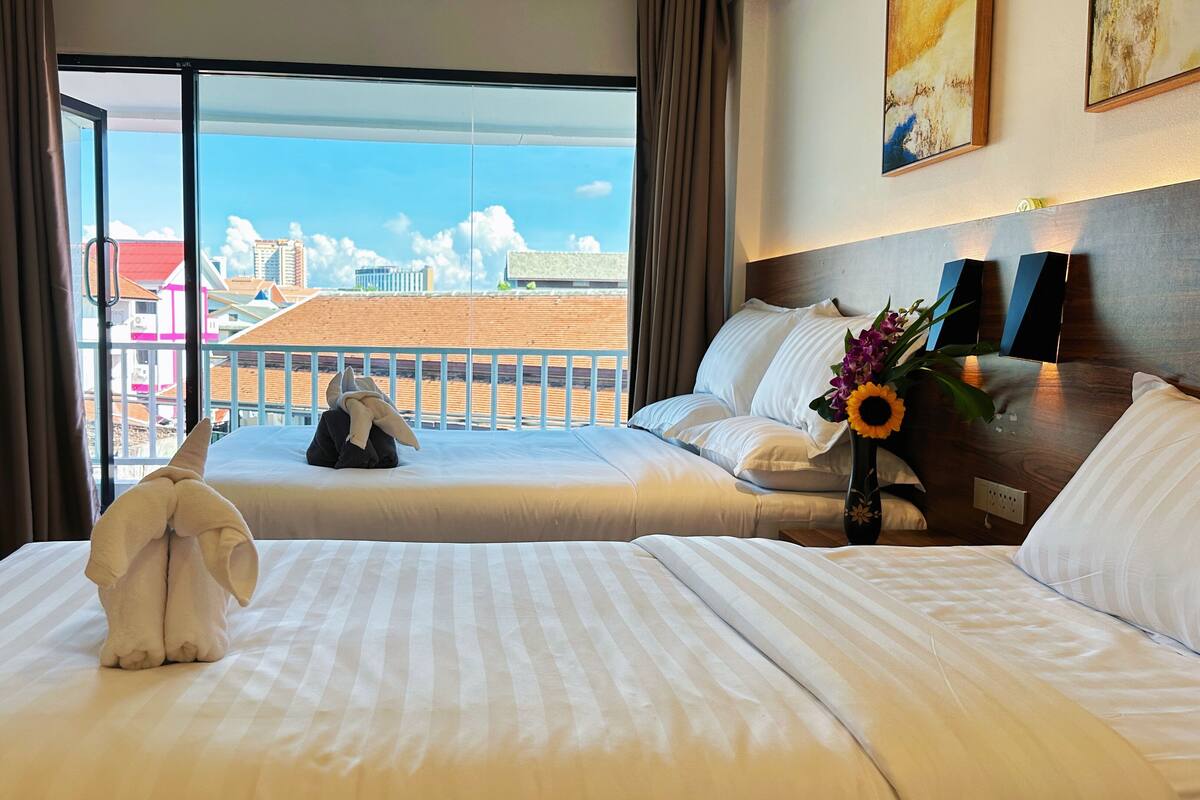 Quarto de hotel com duas camas, uma mesa de trabalho e varanda com vista para edifícios e céu azul.
