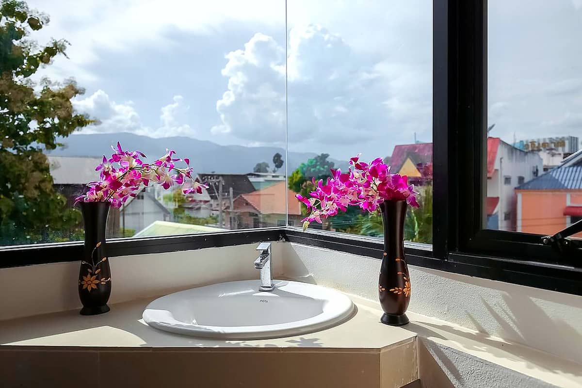 Casa de banho com uma pia e dois vasos de flores num parapeito de janela, com vista para a cidade.