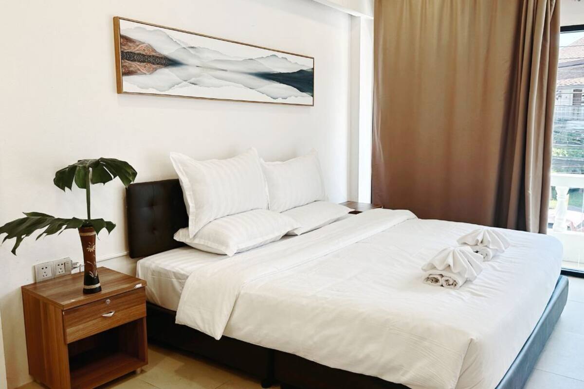 Uma cama bem arrumada com lençóis brancos, uma planta na mesa de cabeceira e um quadro acima.