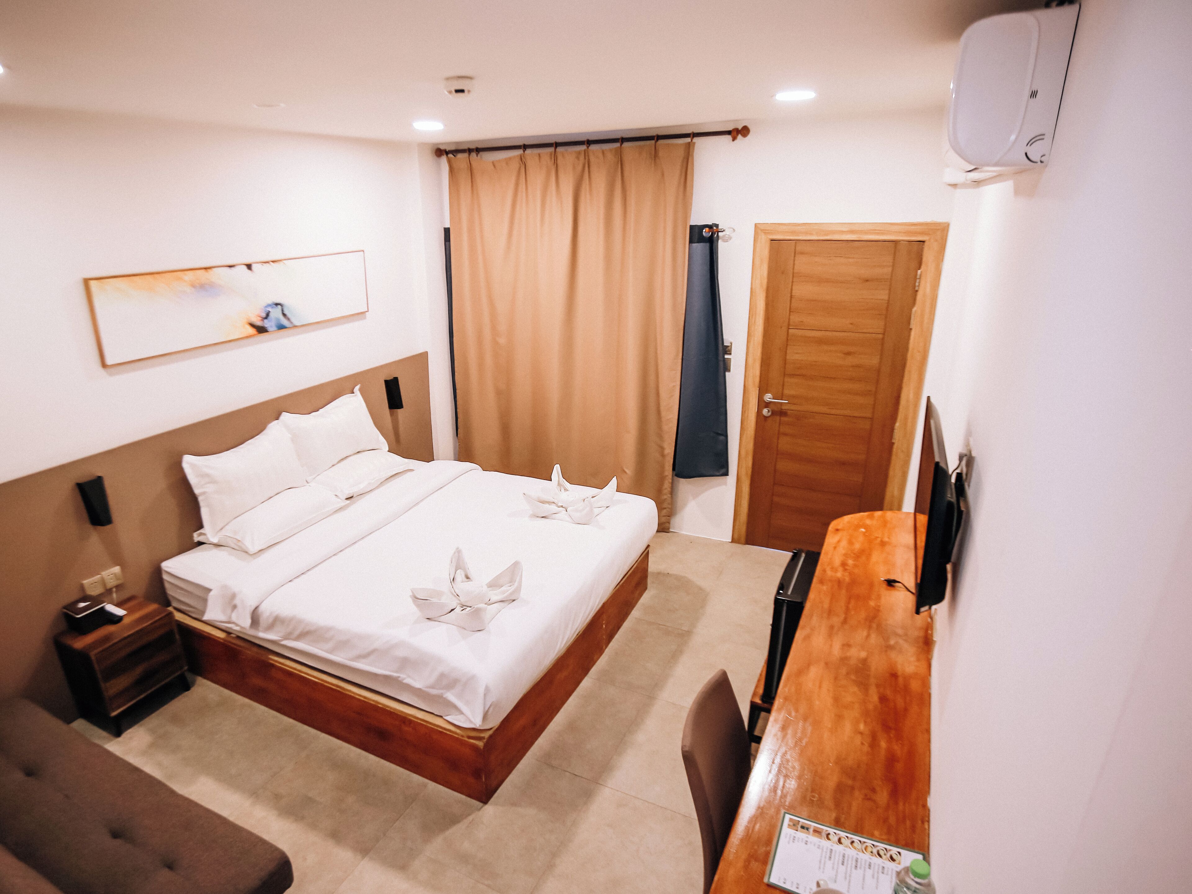 A hotel room with a wooden bed, a desk, a chair, a nightstand, a wall-mounted TV, and a door.