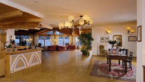 A spacious lobby with a wooden reception desk, comfortable seating, a large chandelier, and a view of the outdoors. - Carlo Magno Hotel Spa Resort (Pinzolo)
