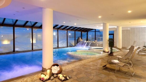A spa area with a large indoor pool, a waterfall feature, and lounge chairs. - Carlo Magno Hotel Spa Resort (Pinzolo)