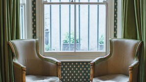 A room with two wooden armchairs facing a window with bars, a red ottoman with books, and a patterned rug. - Glorious 2 Bedroom Apartment (London)
