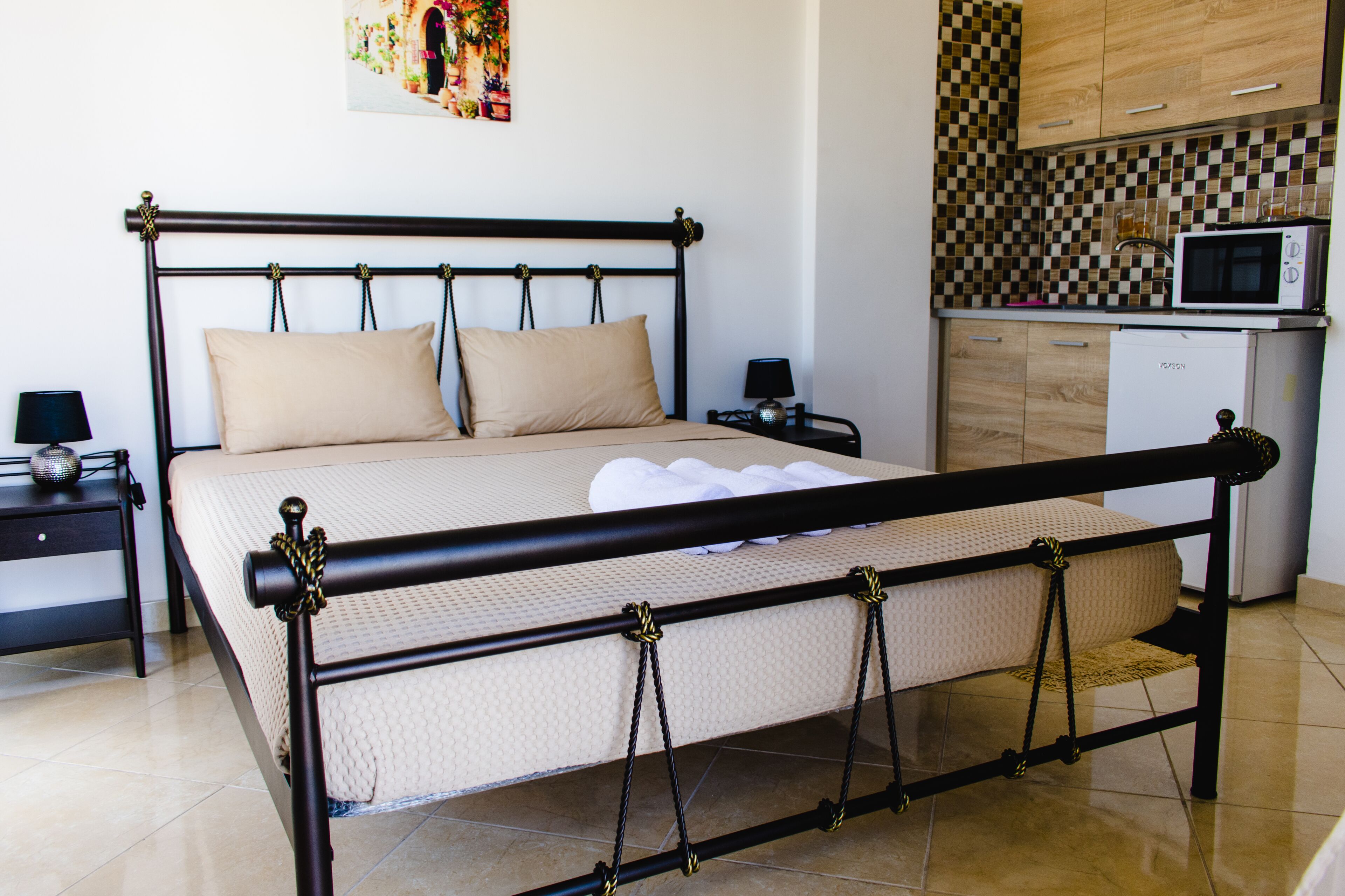 A bedroom with a bed, bedside tables, a nightstand, a microwave, and a kitchen area with cabinets and a tiled backsplash.