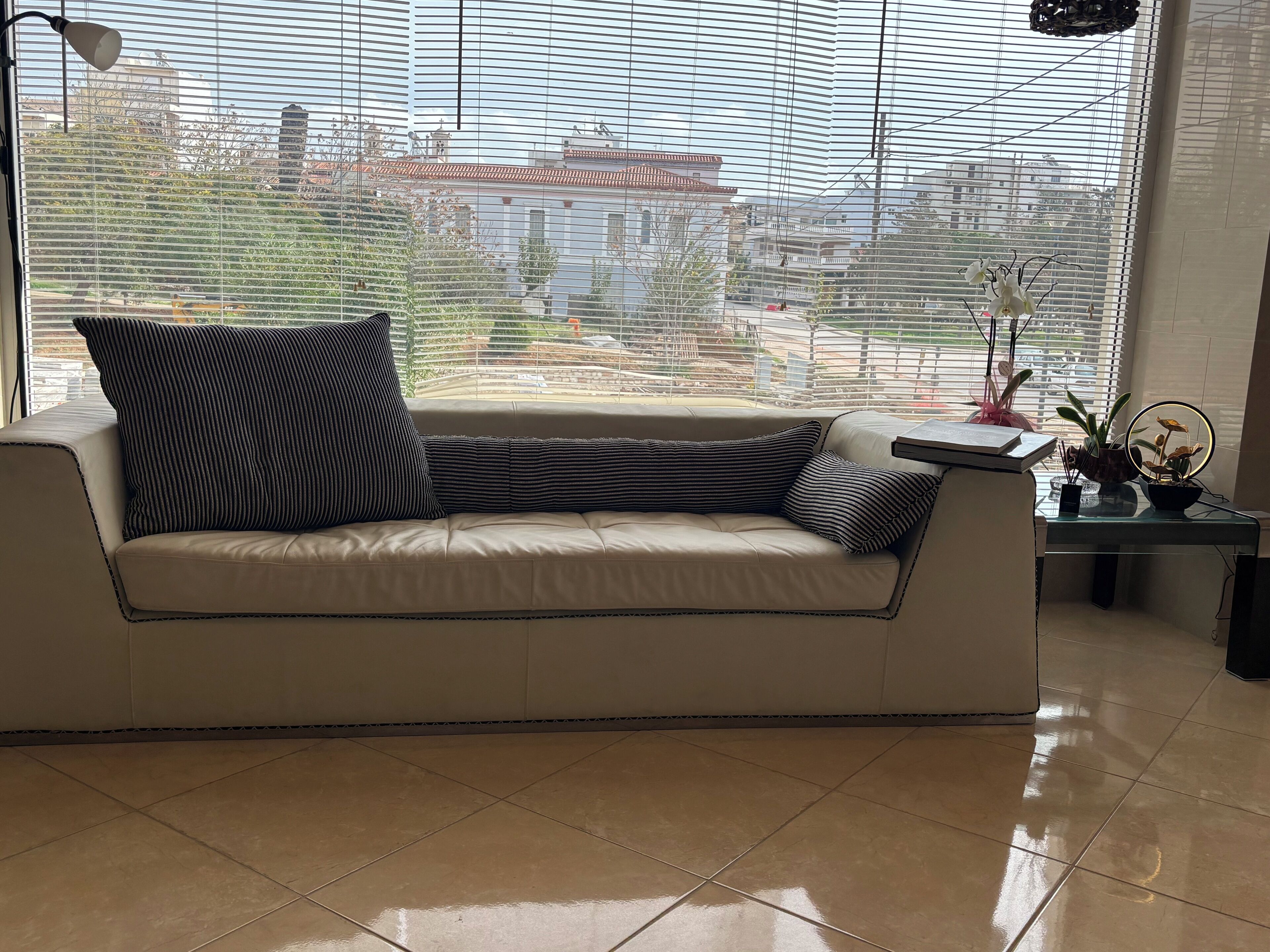 Un sofá color beige con almohadones a rayas, una mesita con una planta y un espejo, y vista a edificios a través de ventanas amplias.