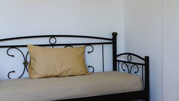 A single bed with a metal frame and a beige mattress, a small black shelf above the bed, and a white towel on a beige mat in the foreground.