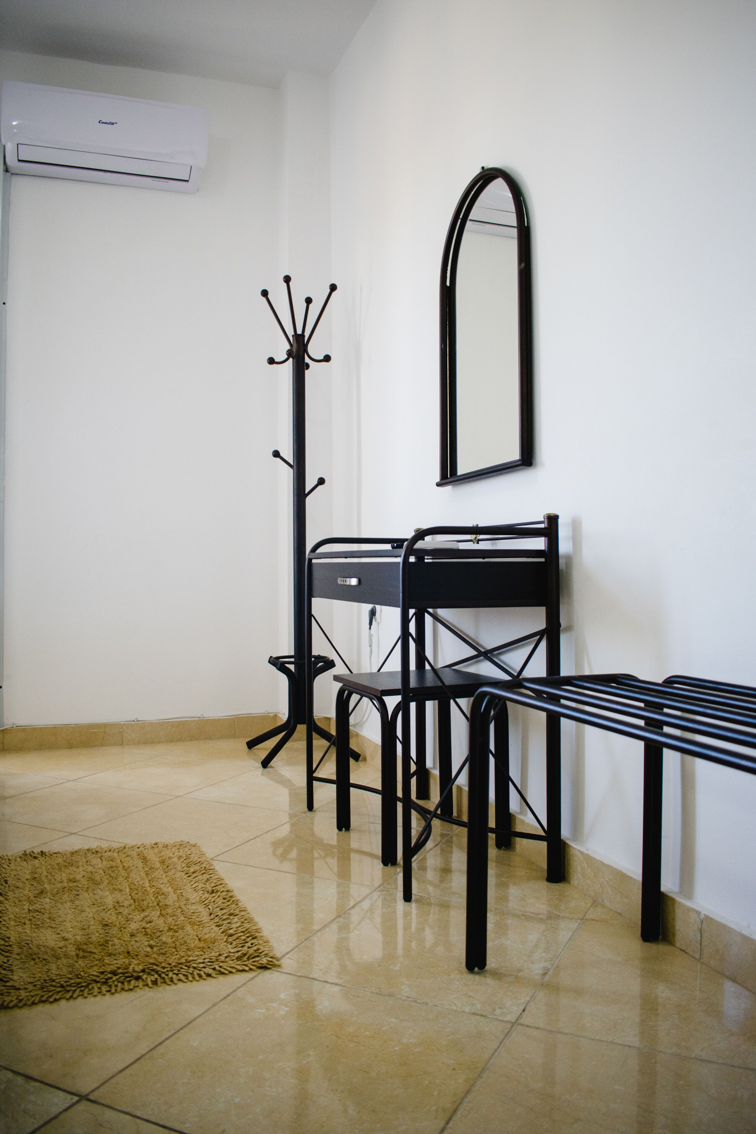 Una habitación con un escritorio de metal negro, un perchero, un espejo y un tapete beige sobre piso de baldosas.