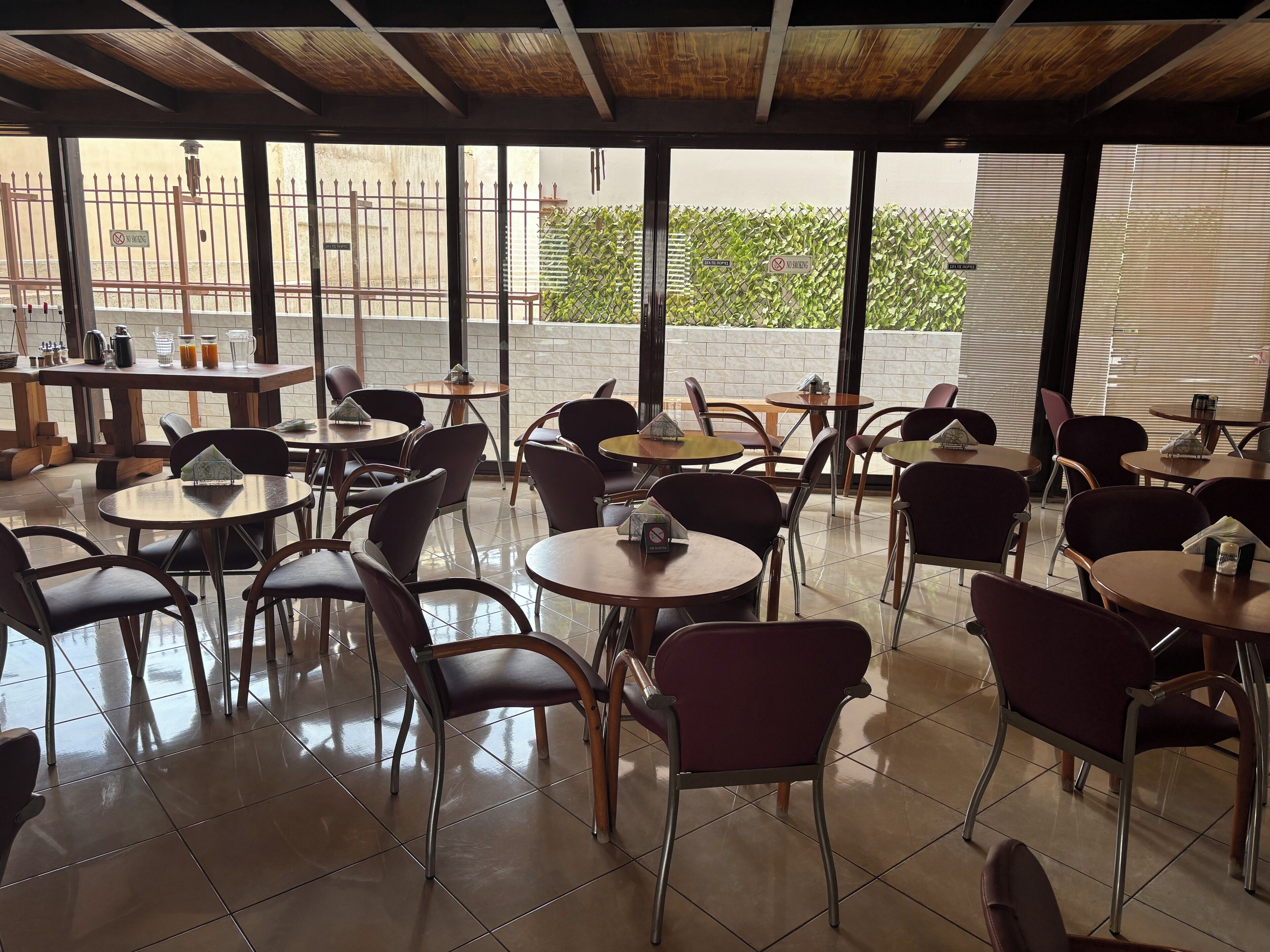 Un restaurante con mesas y sillas redondas, piso de baldosas y amplios ventanales que ofrecen vistas a un edificio y zonas verdes.