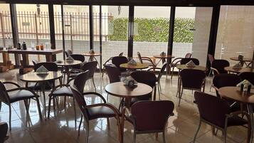 Un restaurante con mesas y sillas redondas, piso de baldosas y amplios ventanales que ofrecen vistas a un edificio y zonas verdes.