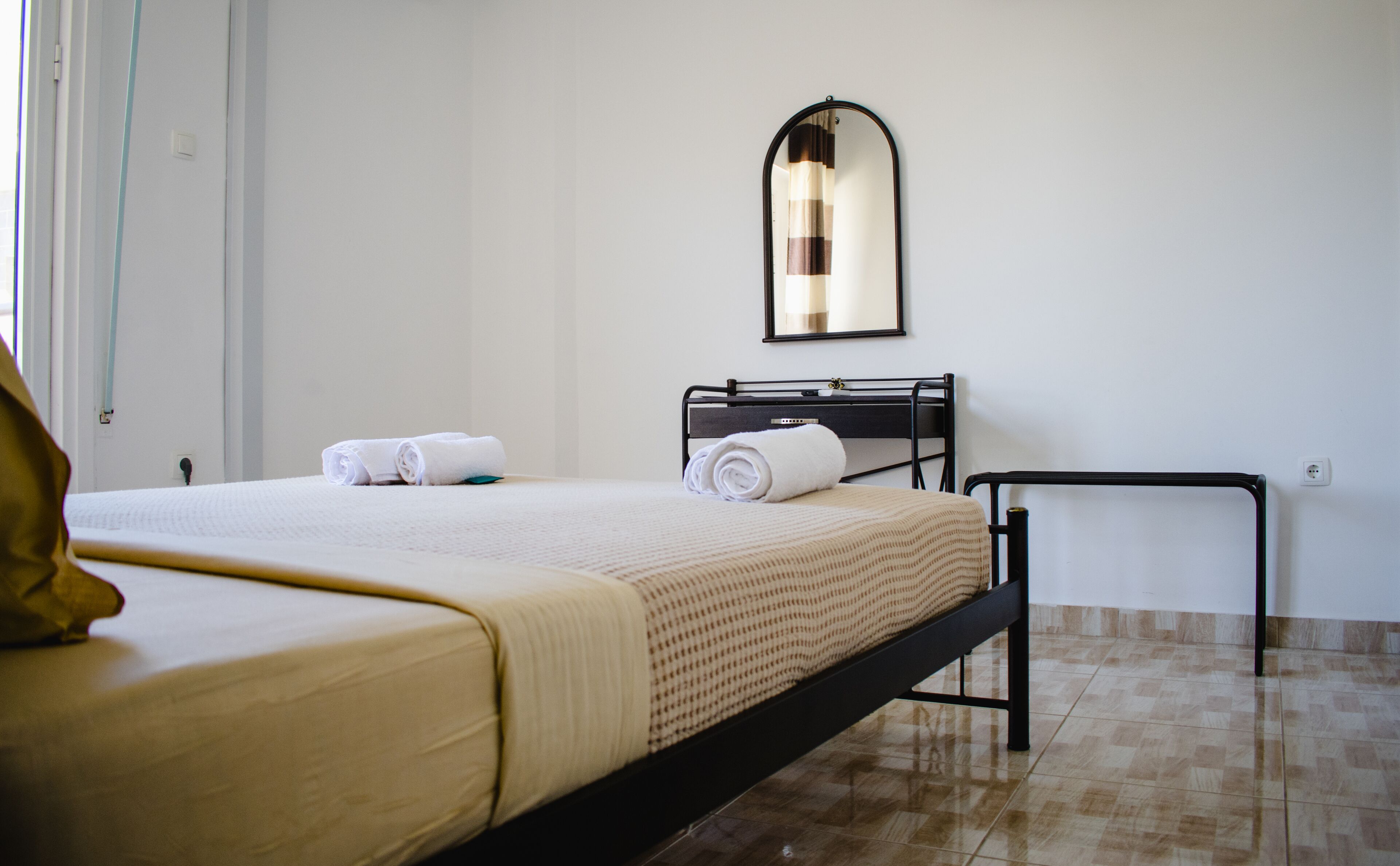Een net opgemaakt bed met een witte deken en een beige plaid, een zwart metalen hoofdbord, een klein nachtkastje en een wandspiegel.