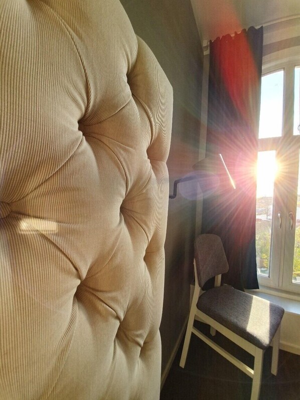 A room with a large, tufted headboard, a small chair, and a window with curtains. - Hotell Carl Friman (Motala)
