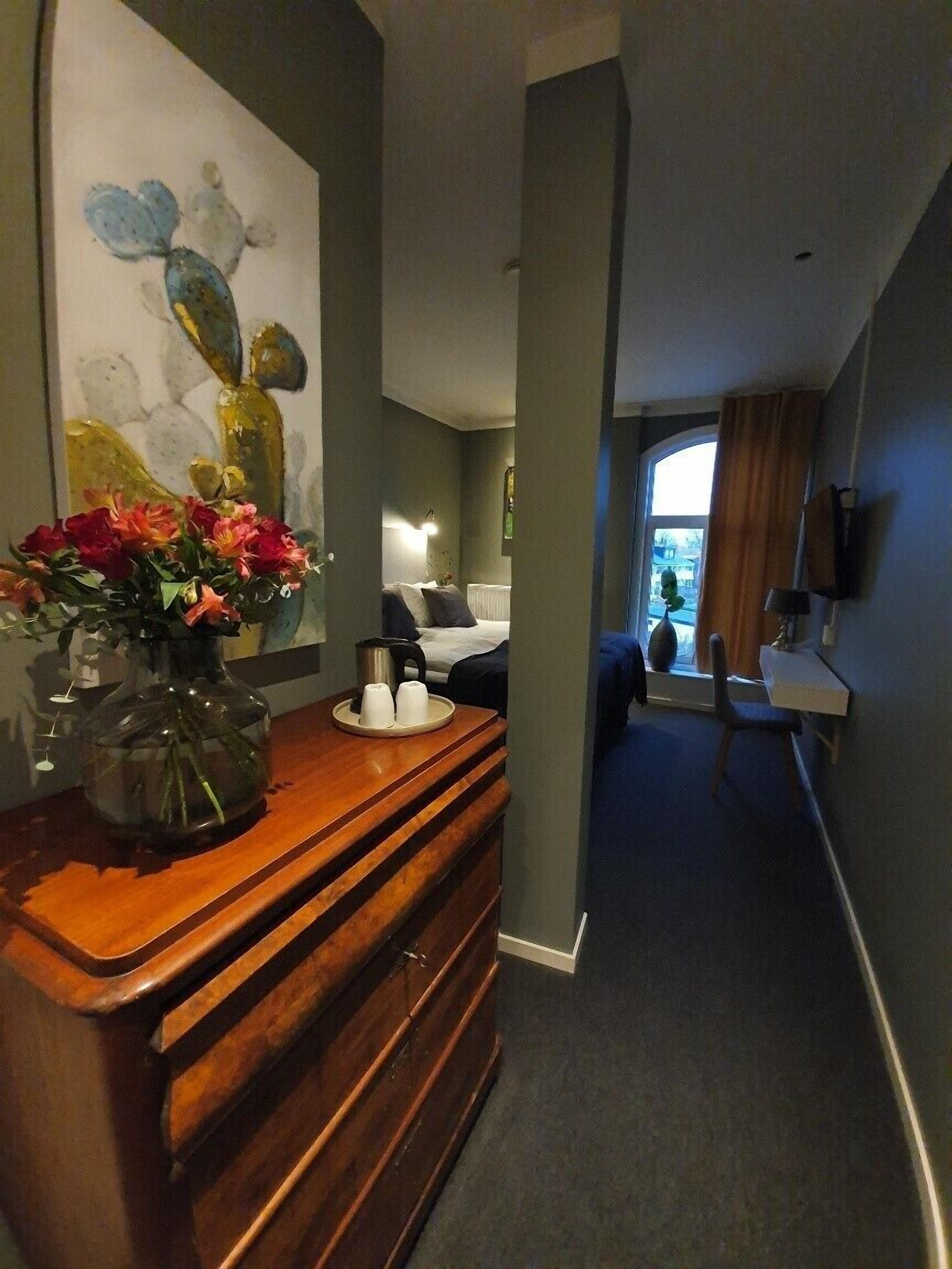 Una habitación de hotel con un aparador de madera, un jarrón de flores, una cama con una mesita de noche, una ventana con cortinas y un cuadro en la pared.