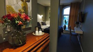 Una habitación de hotel con un aparador de madera, un jarrón de flores, una cama con una mesita de noche, una ventana con cortinas y un cuadro en la pared.