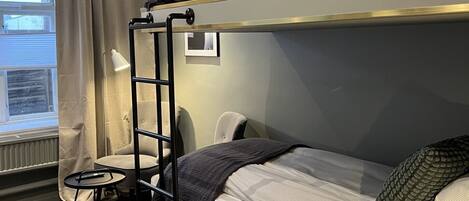 Quarto com beliche, escrivaninha, cadeira, janela com cortinas e radiador.