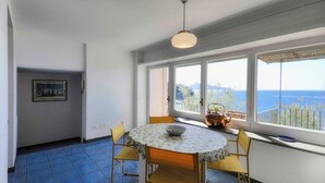 Quarto com mesa redonda, quatro cadeiras, luminária pendente e uma grande janela com vista para o mar.
