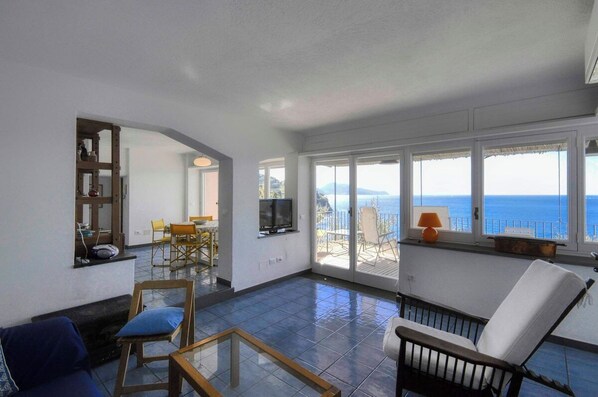 A spacious living area with a large window overlooking the sea, a television, and a dining area with chairs.