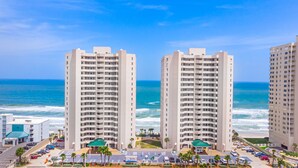 A high-rise hotel with multiple floors, a parking lot, and a view of the ocean. - Oceanfront | Heated Pool-Twin Towers #1405 (Daytona Beach Shores)
