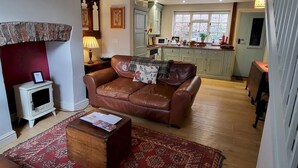 A cozy living room with a brown leather sofa, a small wooden coffee table, and a patterned rug. A kitchen with light green cabinets and a window is visible in the background. - 7 Monk Bar Court YORK (York)