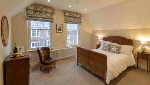 A bedroom with a wooden bed, two windows with floral valances, a radiator, a chair, a bedside table with a lamp, and a dresser with a bottle and glasses. - Apt 2, 71 Micklegate, YORK (York)