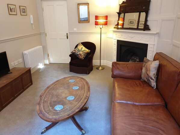 A living room with a brown leather sofa, a wooden coffee table with decorative plates, a dark armchair with a floral cushion, a standing floor lamp, a white fireplace, and a white door. - Apt 1, 71 Micklegate, YORK (York)