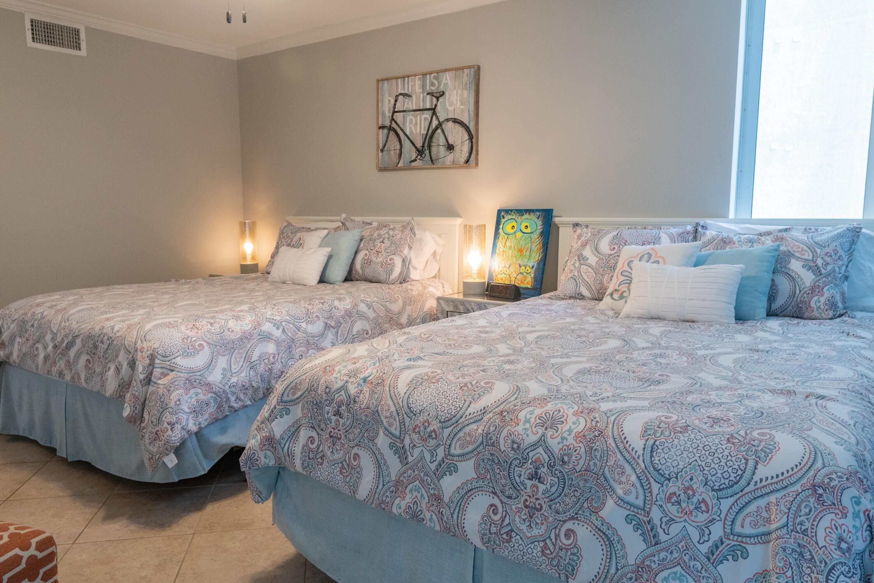 A bedroom with two beds, patterned bedding, wall art, and bedside lamps.