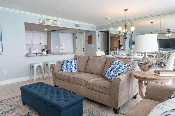 Una sala de estar con un sofá color beige, cojines estampados azules, un reposapiés azul y una mesita redonda con un libro y un cuenco de vidrio.
