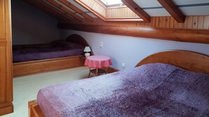 Une chambre avec un plafond en pente, des poutres en bois, un puits de lumière, deux lits, une petite table recouverte d’une nappe rouge et blanche, et une couverture violette.