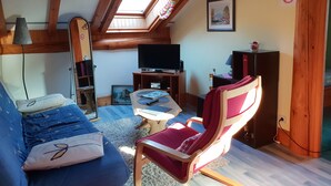 Un salon chaleureux avec un plafond en pente, des poutres en bois, un canapé bleu, un fauteuil rouge, une télévision et une petite table.