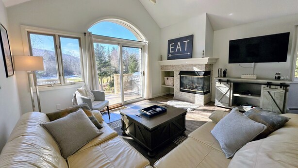 A spacious living room with a fireplace, a television, and a large window offering a view of the outdoors.