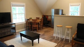 Una sala de estar acogedora con televisor, muebles de madera y una zona de cocina con refrigerador y taburetes de bar.
