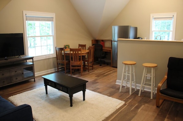 A cozy living room with a TV, wooden furniture, and a kitchen area with a refrigerator and bar stools.