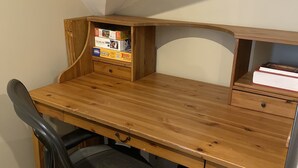 Un escritorio de madera con una silla, un estante pequeño con cajas de acertijos, y un libro a la derecha.