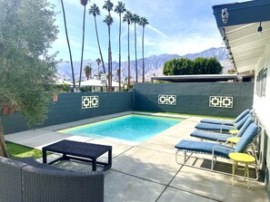 A swimming pool with lounge chairs and a table, surrounded by a low wall with decorative patterns, and palm trees in the background. - Casa Sunny Dunes: a Private, Mid-Century Oasis (Palm Springs)