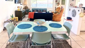 Un comedor con una mesa dispuesta para cuatro, rodeada de sillas verdes, un sofá y un televisor al fondo.