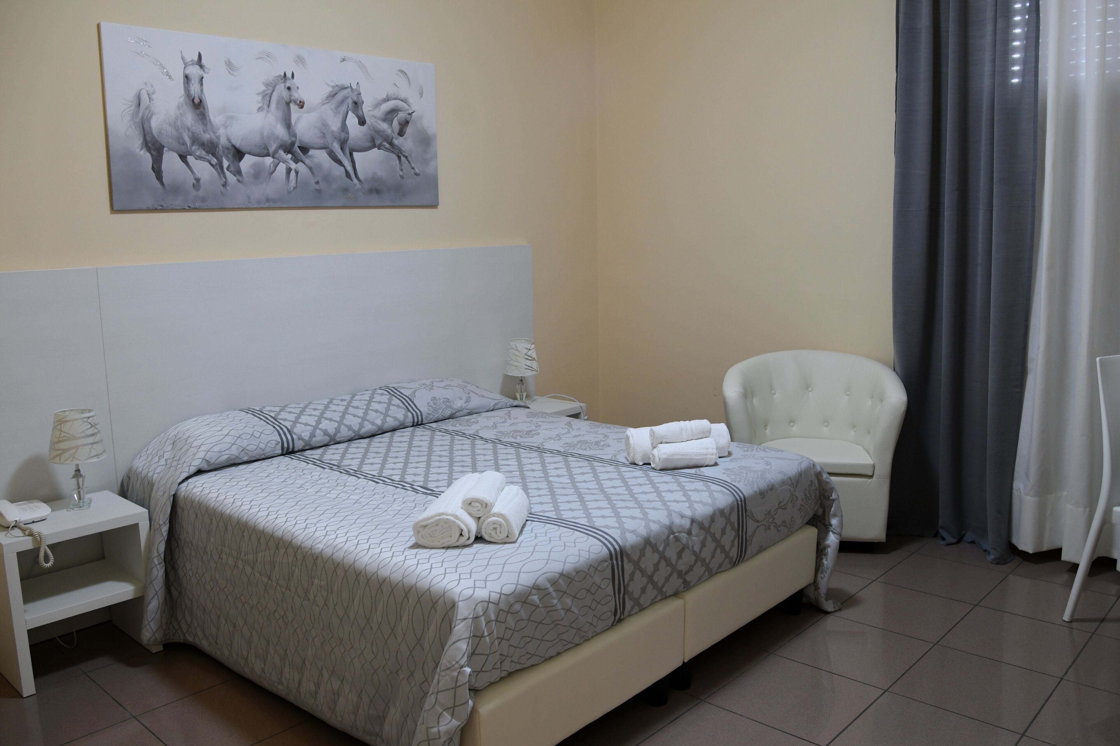 Un dormitorio ordenado con una cama grande, una mesita de noche con lámpara, una silla y un cuadro de caballos en la pared.