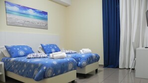 A hotel room with two beds, each with blue bedding and white pillows, a nightstand, a small table, and a painting of a beach scene on the wall. - Hotel Villa Romana (Piazza Armerina)
